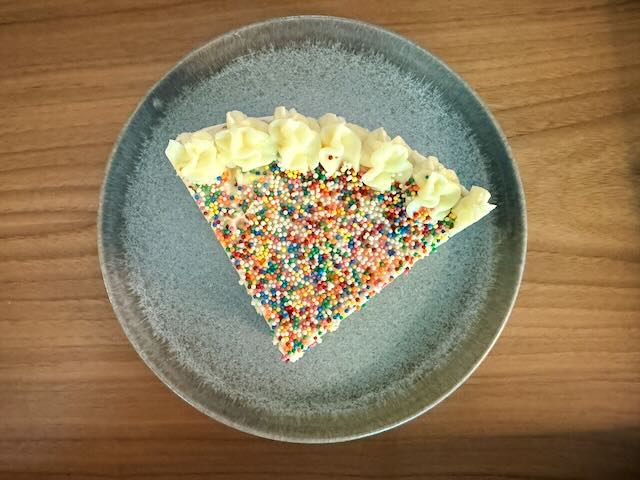 Triangular cake slice topped with rainbow sprinkles and piped pale yellow frosting on a blue-grey plate
