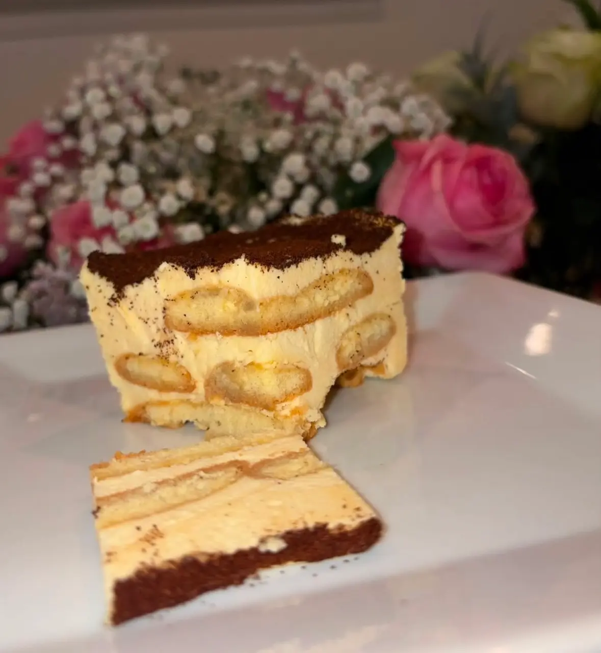Cocoa-dusted tiramisu slice with ladyfinger layers on a white plate, pink roses behind